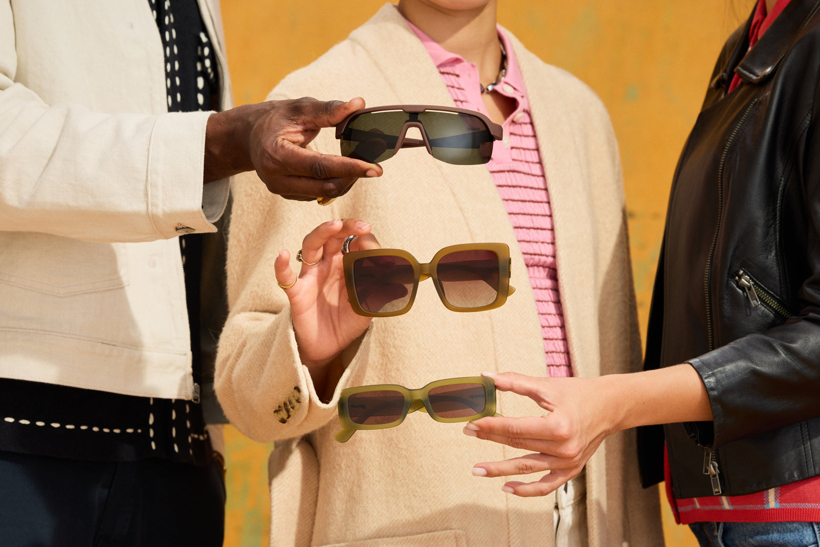 Three people holding different styles of goodr sunglasses in front of a warm yellow wall. The sunglasses feature olive green frames and dark lenses, shown in a lifestyle setting.