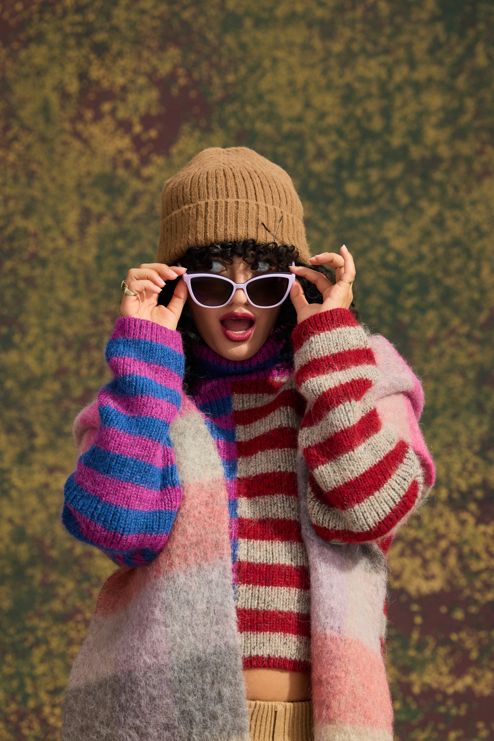 A person wearing a tan knit beanie, purple Pounce G goodr cat-eye sunglasses, a pink, purple, blue, and gray striped knit sweater vest over a red and white striped knit top, posed against a textured olive green background.
