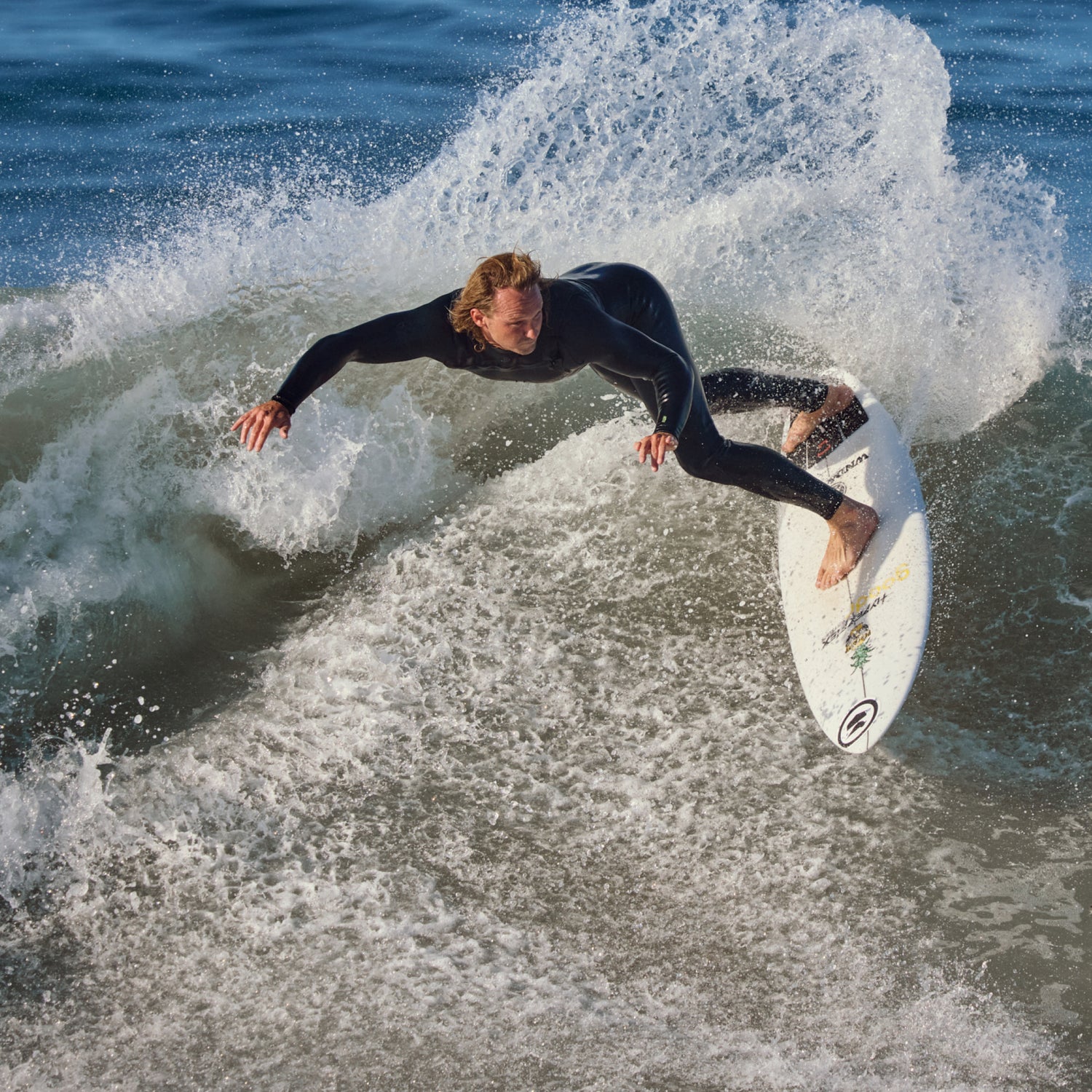 A surfer in a black wetsuit rides a white surfboard on a breaking ocean wave, with white spray and blue water around them.