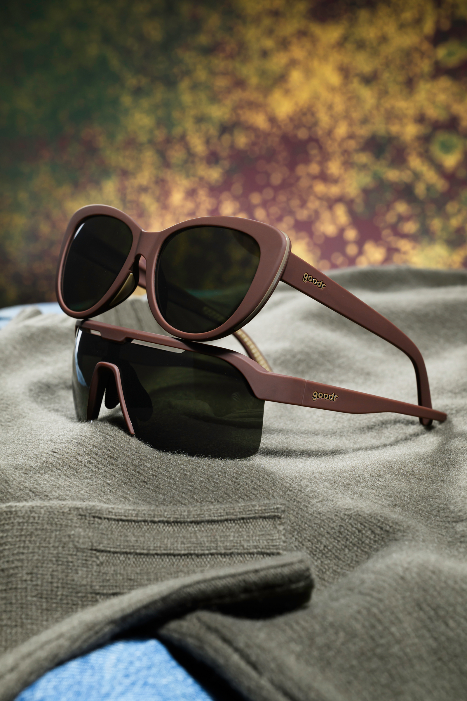 Two pairs of goodr sunglasses with matte brown frames and dark lenses, stacked on a gray knit sweater with a blurred autumnal background.