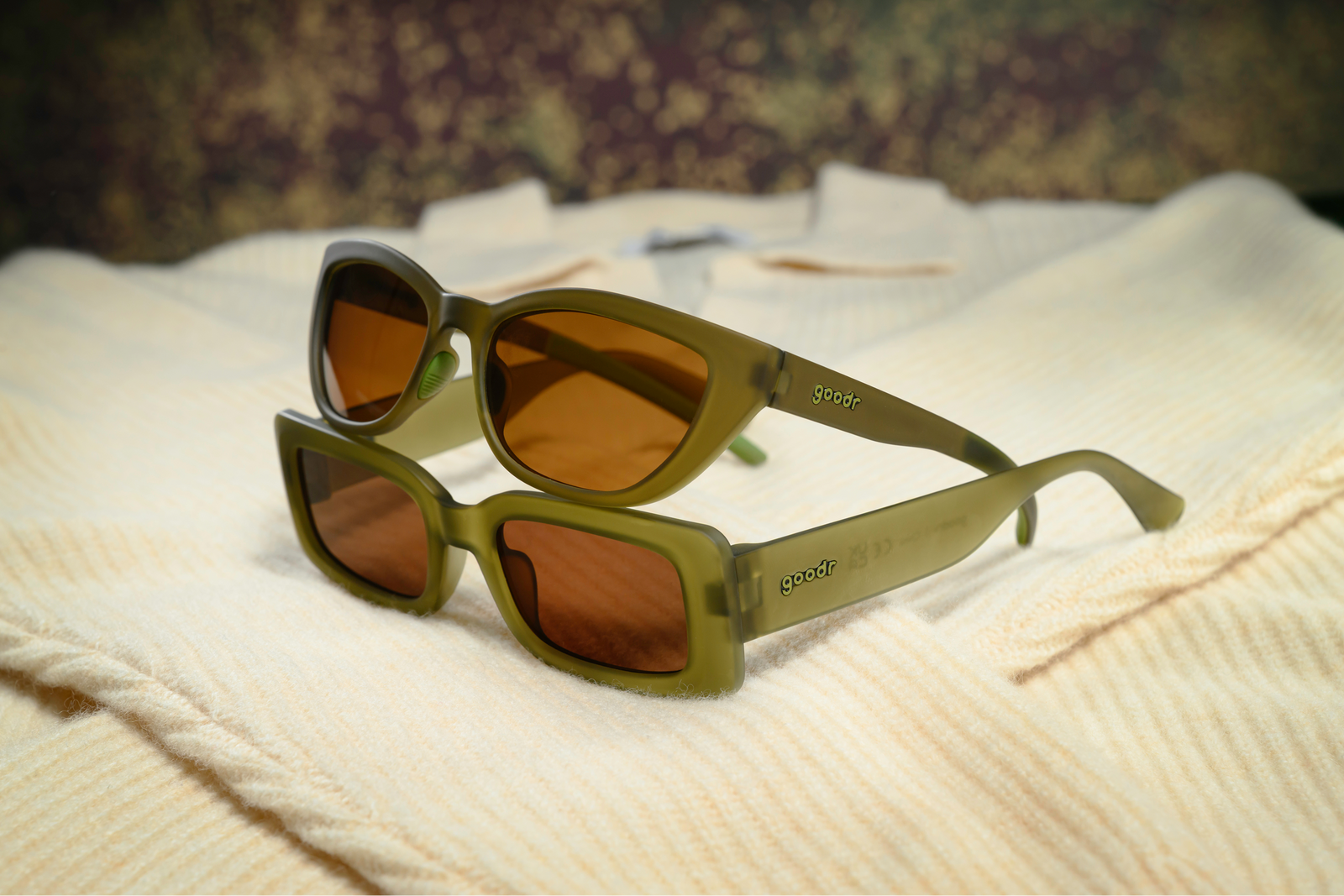 Two pairs of goodr sunglasses with olive green frames and amber lenses, resting on a cream-colored ribbed knit fabric.