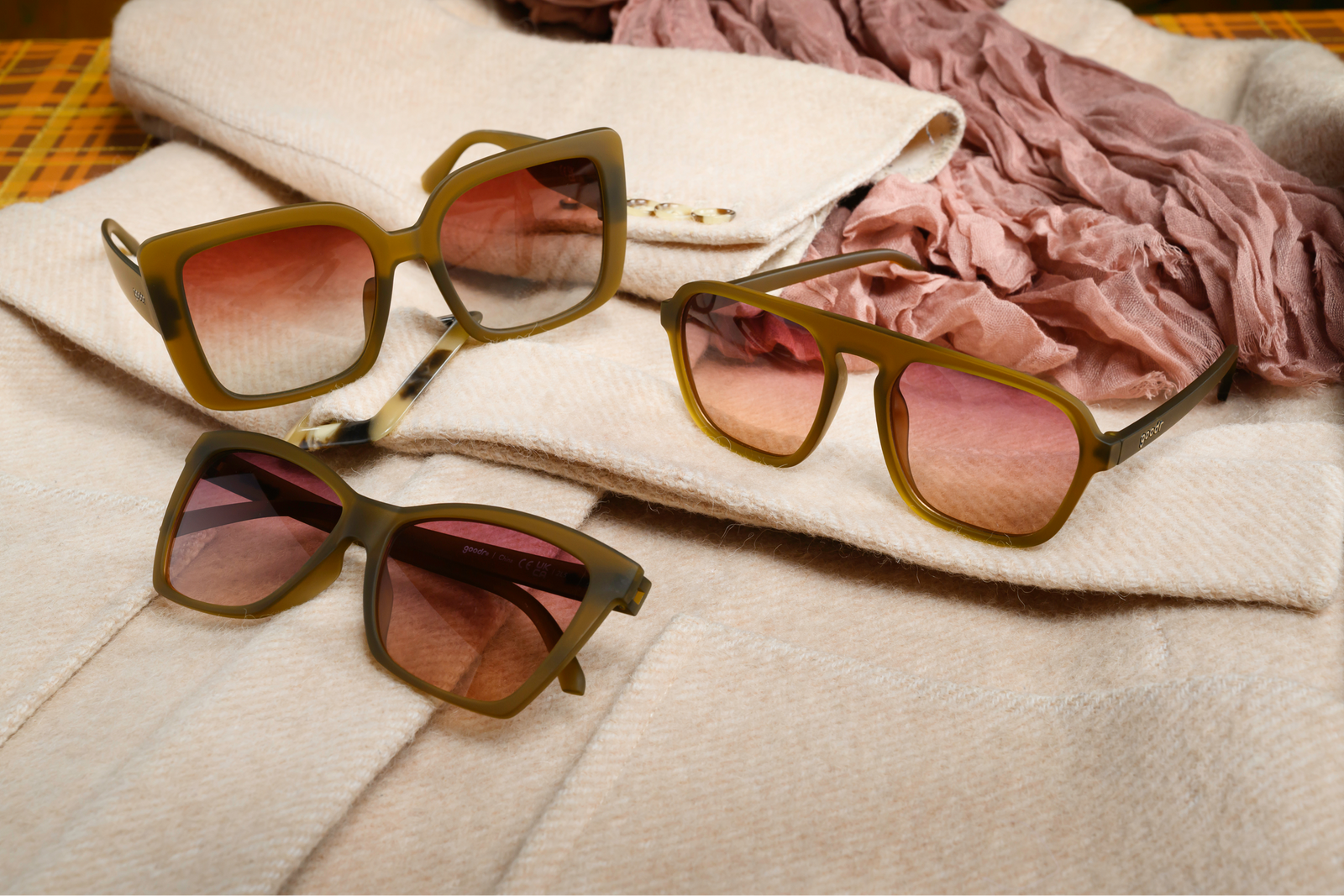 Three pairs of goodr sunglasses with olive green frames and pink gradient lenses, styled on a cream-colored tweed jacket with a dusty pink scarf.
