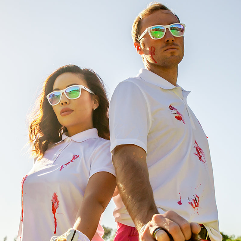 A man and woman in white sunglasses with rose lenses confidently stand back-to-back, blood spatter on their golf shirts.
