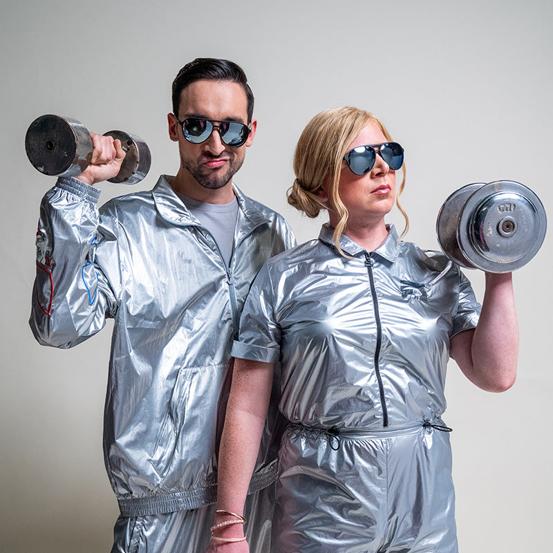 A man and woman wearing black and chrome aviator sunglasses and silver jumpsuits hold barbells with fierce facial expressions.