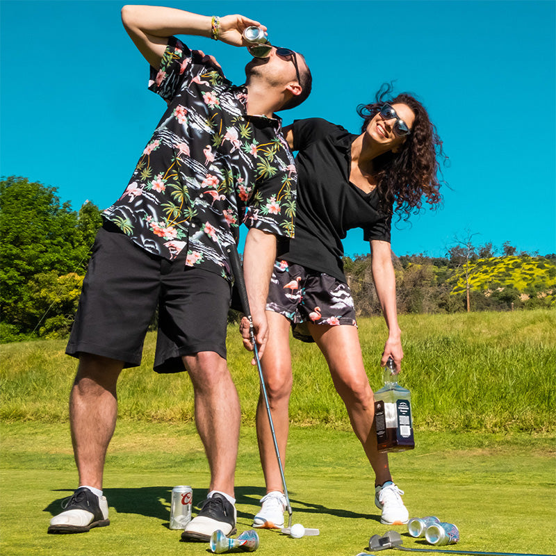 A man and woman wearing black sunglasses stand on a golf course surrounded by beer and liquor while the man shotguns a beer.