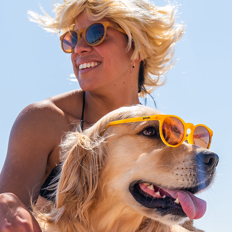 A smiling woman in a blonde wig and a golden retriever wear wood grain patterned circle sunglasses with brown round lenses.