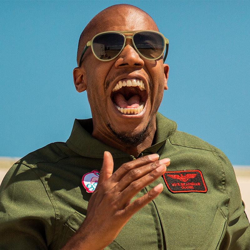 A man in a green military-style jumpsuit and cadet green aviator sunglasses with green gradient lenses shouts joyfully.
