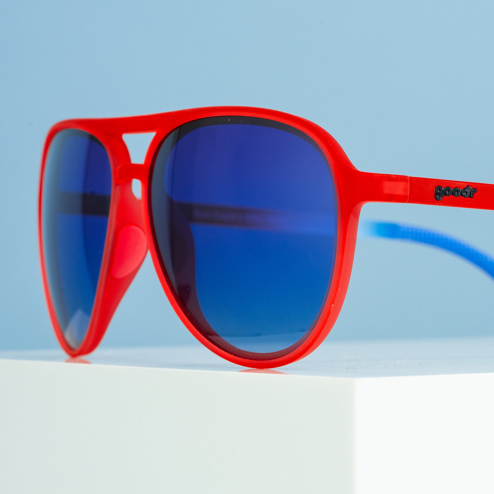 Three-quarter angle close-up view of red, white,+blue aviator sunglasses with blue gradient lenses on a blue background.