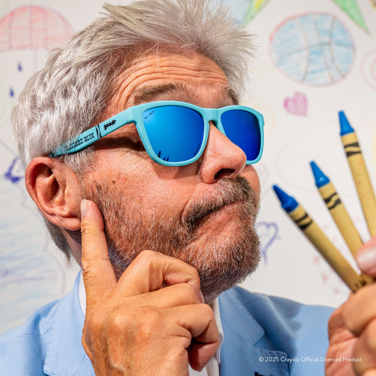 An amused gray-haired man wears Blizzard Blue Crayola sunglasses in front of a white background with colorful drawings