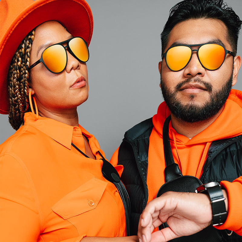 A woman and a man in orange & black sunglasses with mirrored lenses and aviator-style frames vibe as he checks his watch.