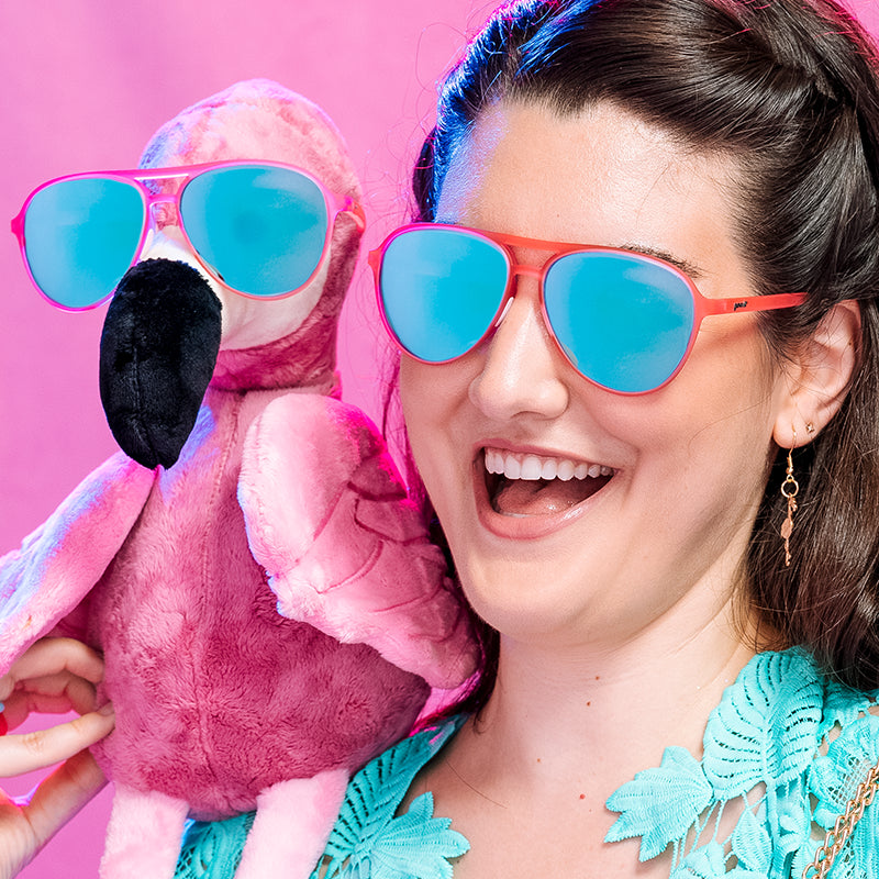 A woman wearing matching pink aviator sunglasses with mirrored reflective teal lenses with her best stuffed flamingo friend.