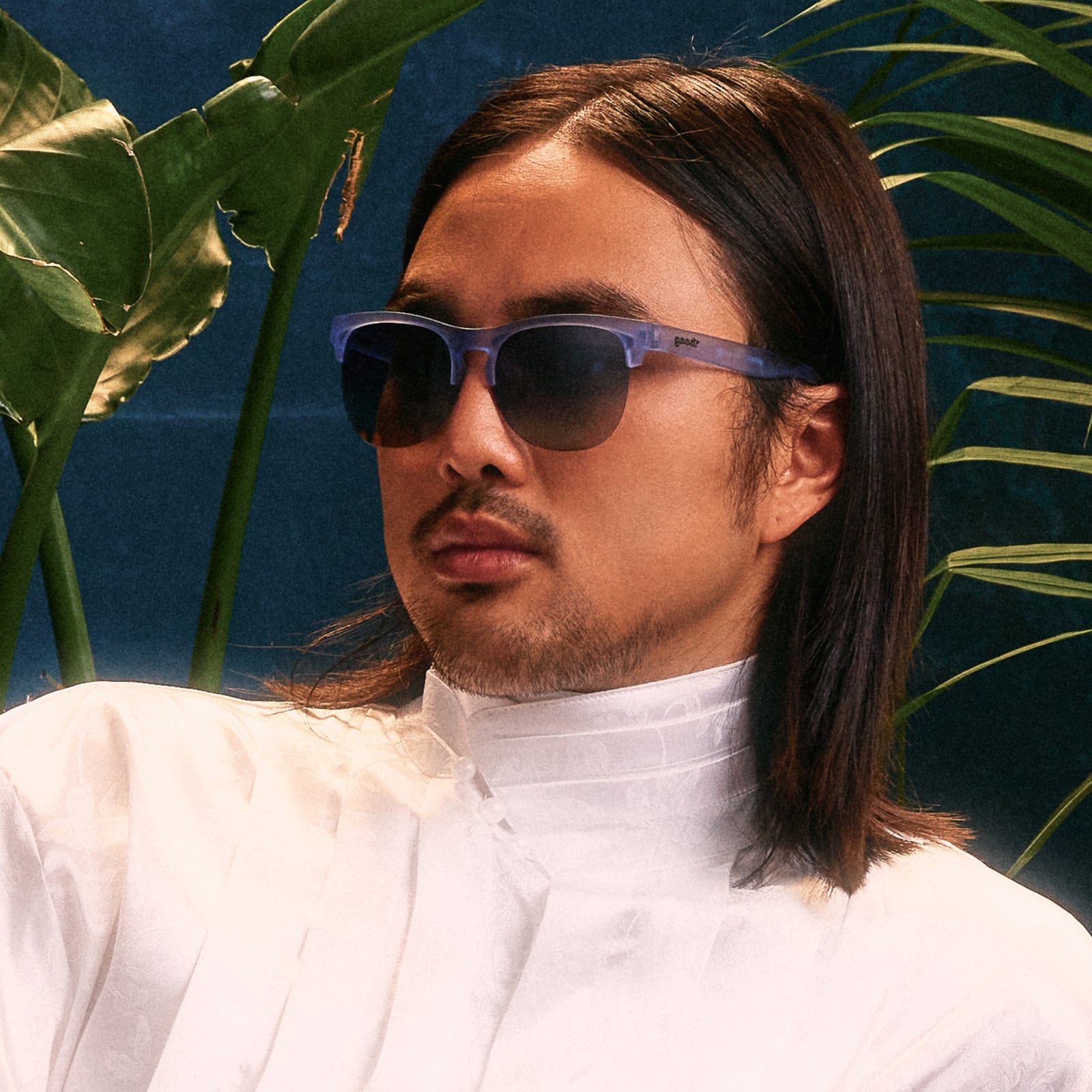 An elegant young man with shoulder-length hair poses in a pair of dusty blue translucent Avant Gs against a blue backdrop.