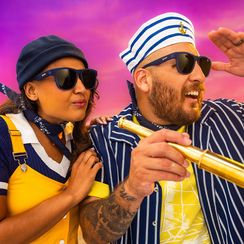 A man and woman in navy blue sunglasses with grey yellow gradient lenses and matching sailor gear watch a beautiful ocean sunset.