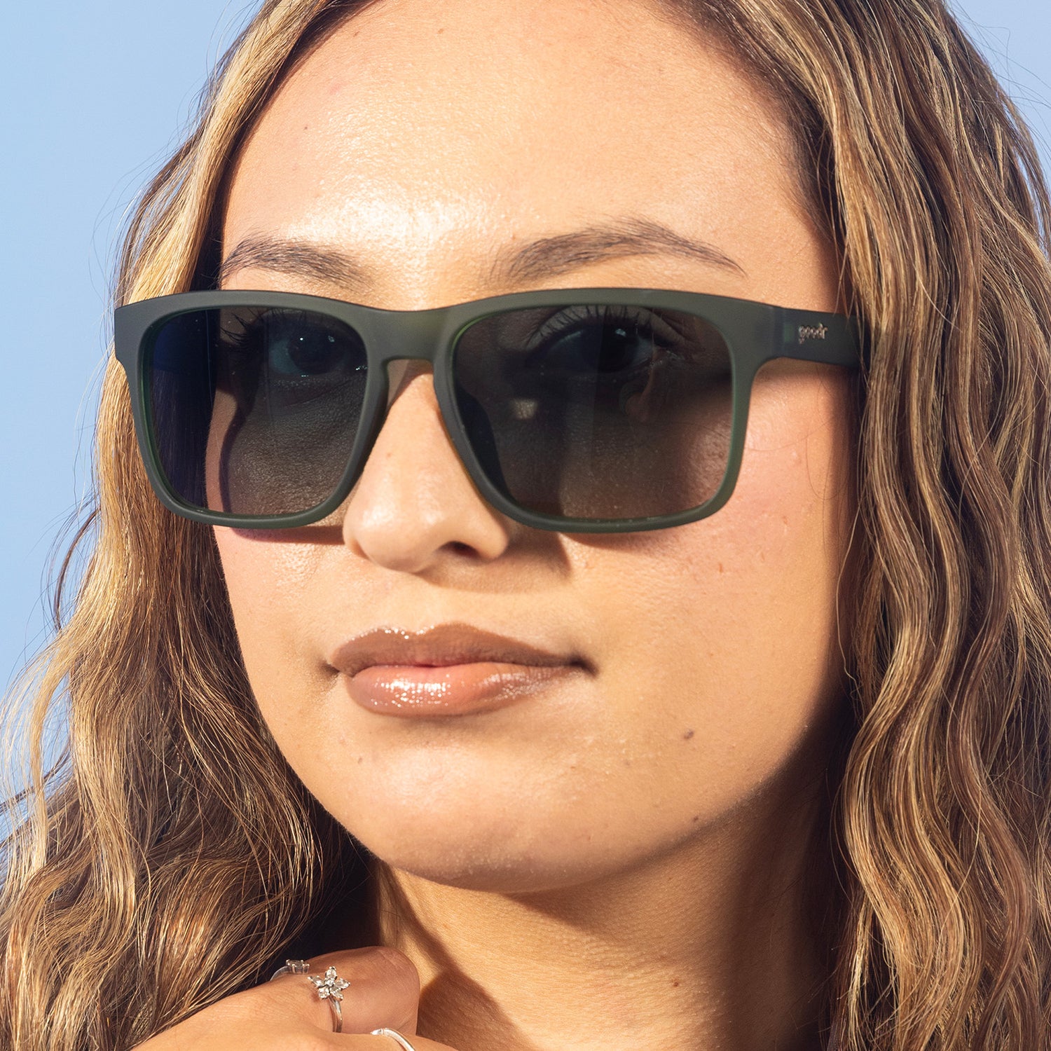 A stylish woman with blonde wavy hair smirks at the camera in translucent dark green Grand Gs with green gradient lenses.