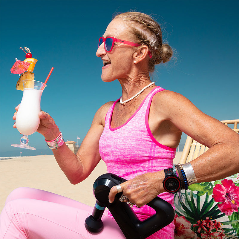A female runner in hot pink sunglasses with green lenses lounges on a beach with a piña colada and massage gun for her legs.