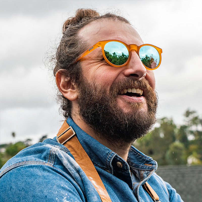 A bearded man with a man bun smiles wearing round orange sunglasses with light blue lenses and an apron covered in flour.