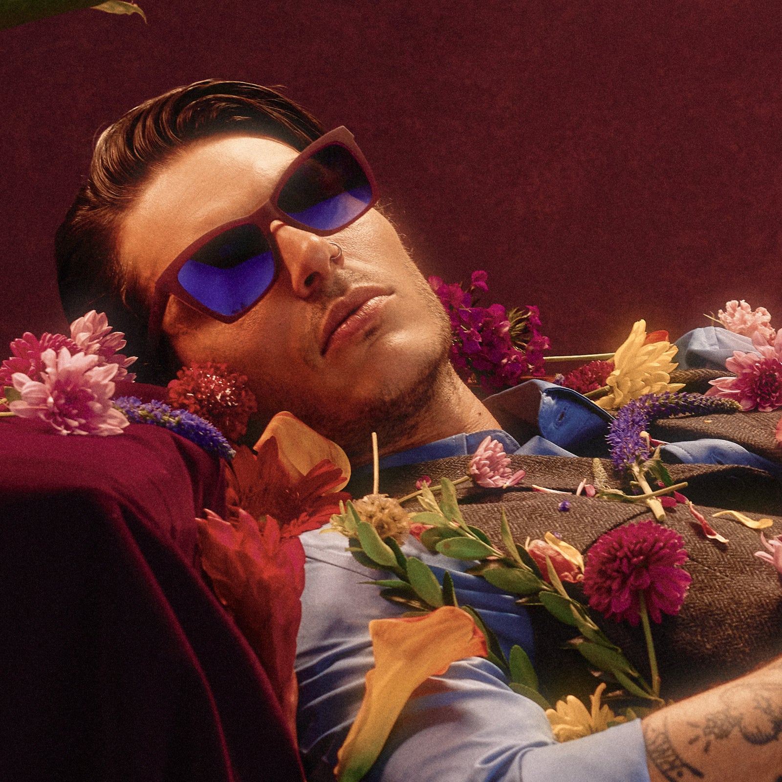 A handsome young man in angular violet sunglasses reclines in a moody space, a variety of flowers is scattered around him.