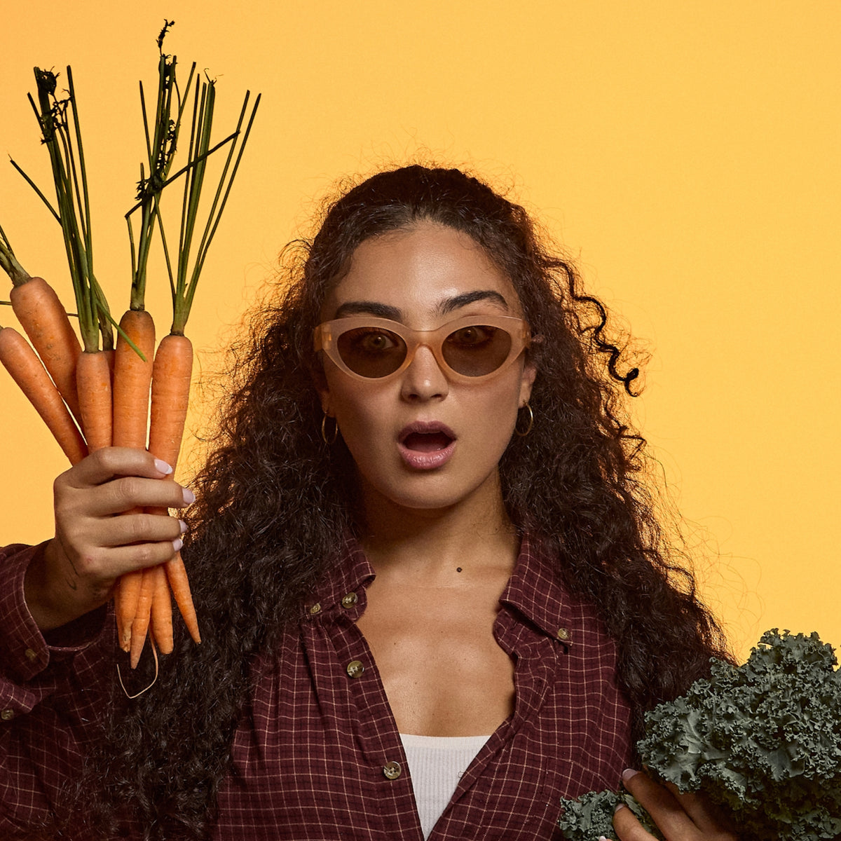 A surprised woman wearing translucent peach cat-eye sunglasses by goodr holds vegetables.