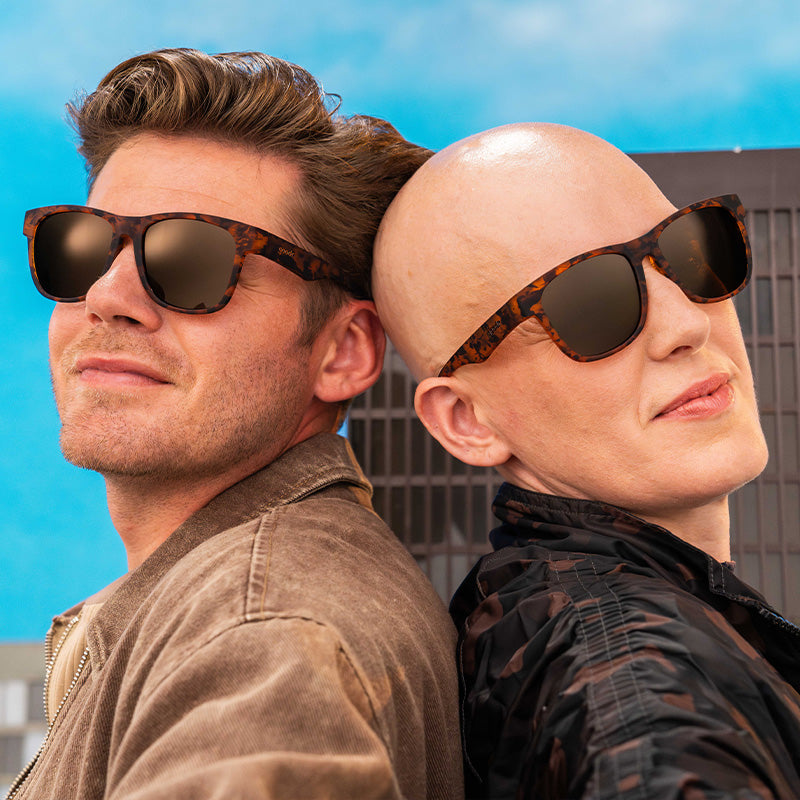 Two people in large tortoiseshell sunglasses with brown non-reflective stand back-to-back before a miniature skyscraper.