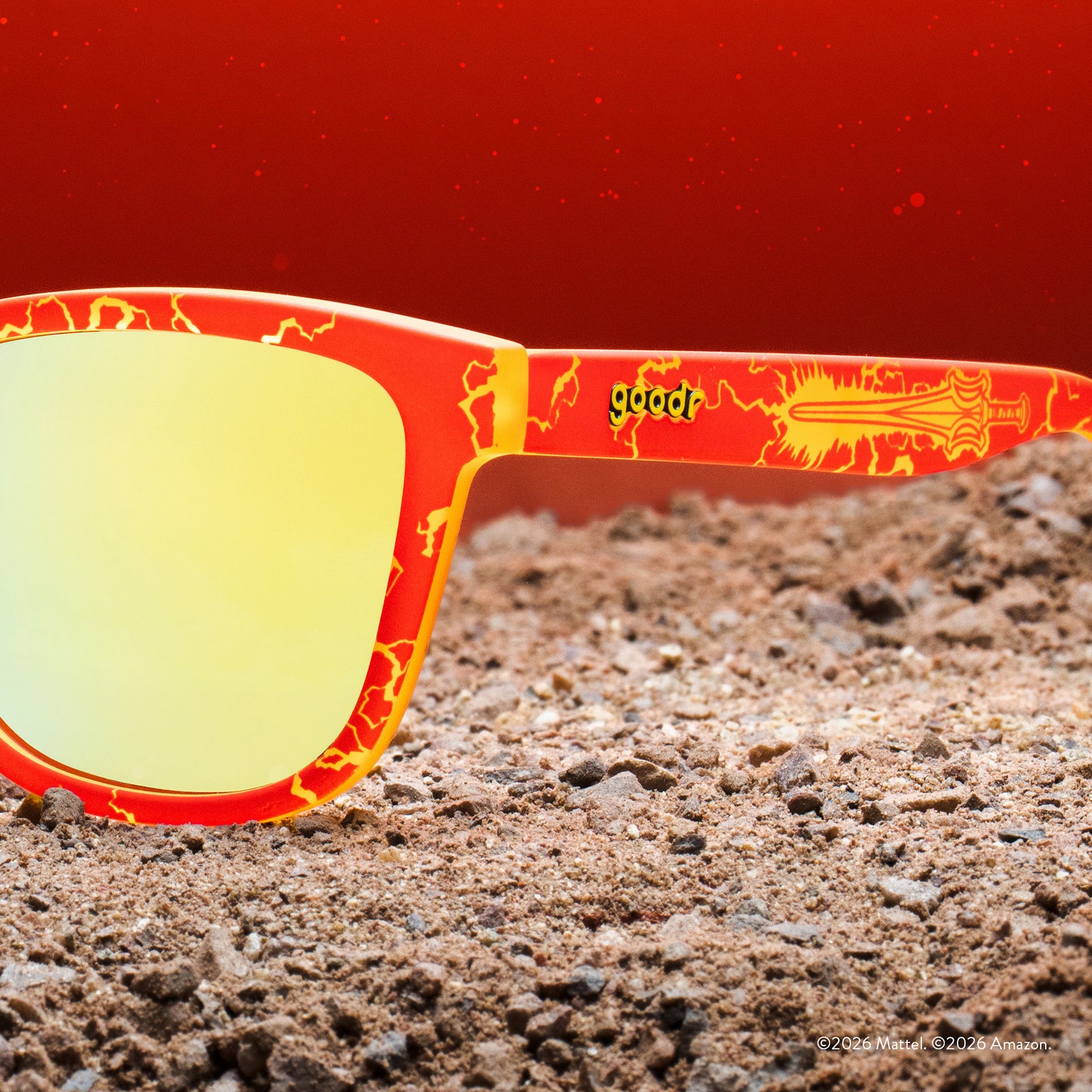 Close-up of the lightning bolt-patterned sunglasses featuring the Masters of the Universe power sword, set against a red, gravel & dirt backdrop.