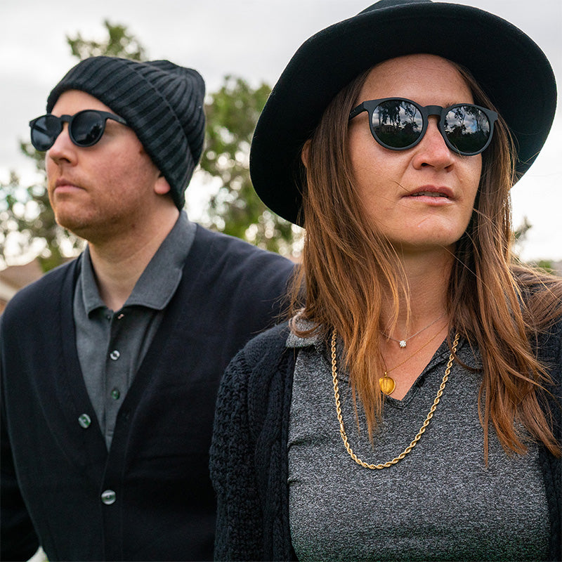 A hipster man and woman wearing chic black outfits wear round black sunglasses with non-reflective black lenses.