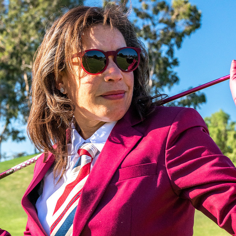 A woman in a dark red power suit and round burgundy sunglasses with brown lenses slings a golf club over her shoulder.