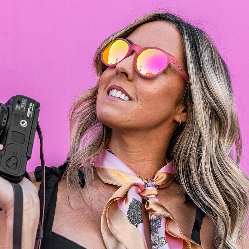A stylish woman wearing round pink sunglasses and holding a camera looks off to the side, a hot pink wall behind her.