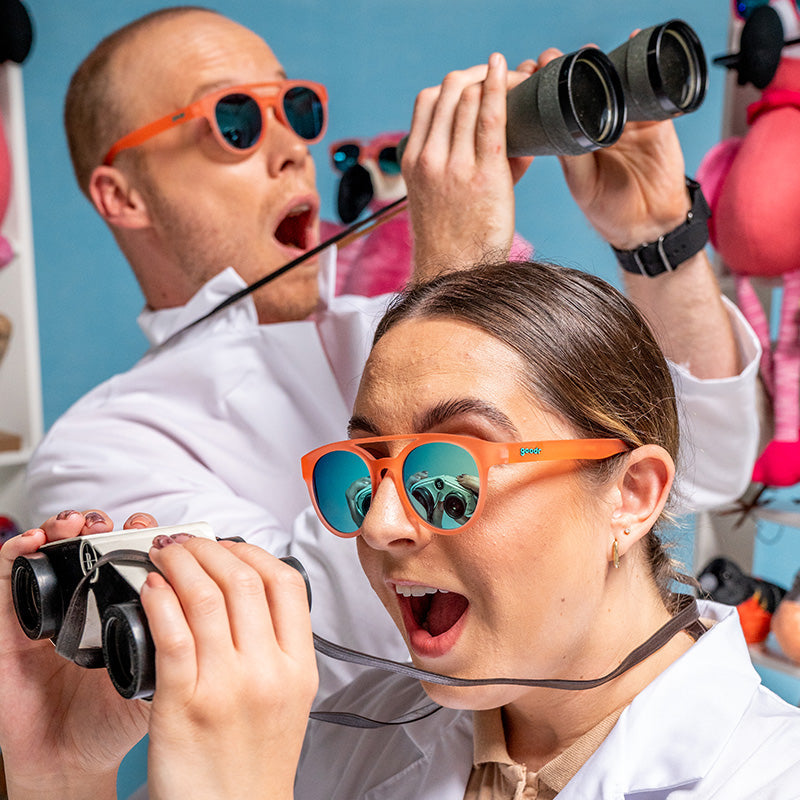 Two joyfully stunned bird watchers in round orange sunglasses with blue reflective lenses gawk through their binoculars.