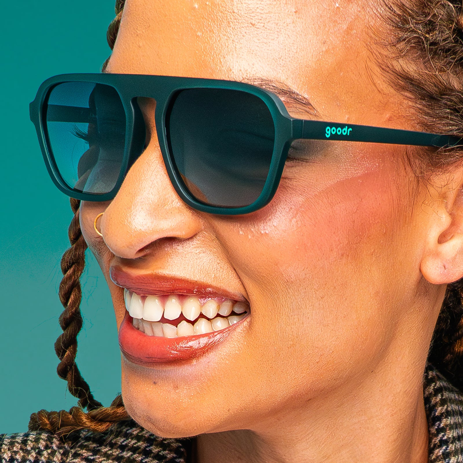 A woman with copper twist braids in a houndstooth coat and translucent teal Retro Gs with green gradient lenses smiles against a teal backdrop.