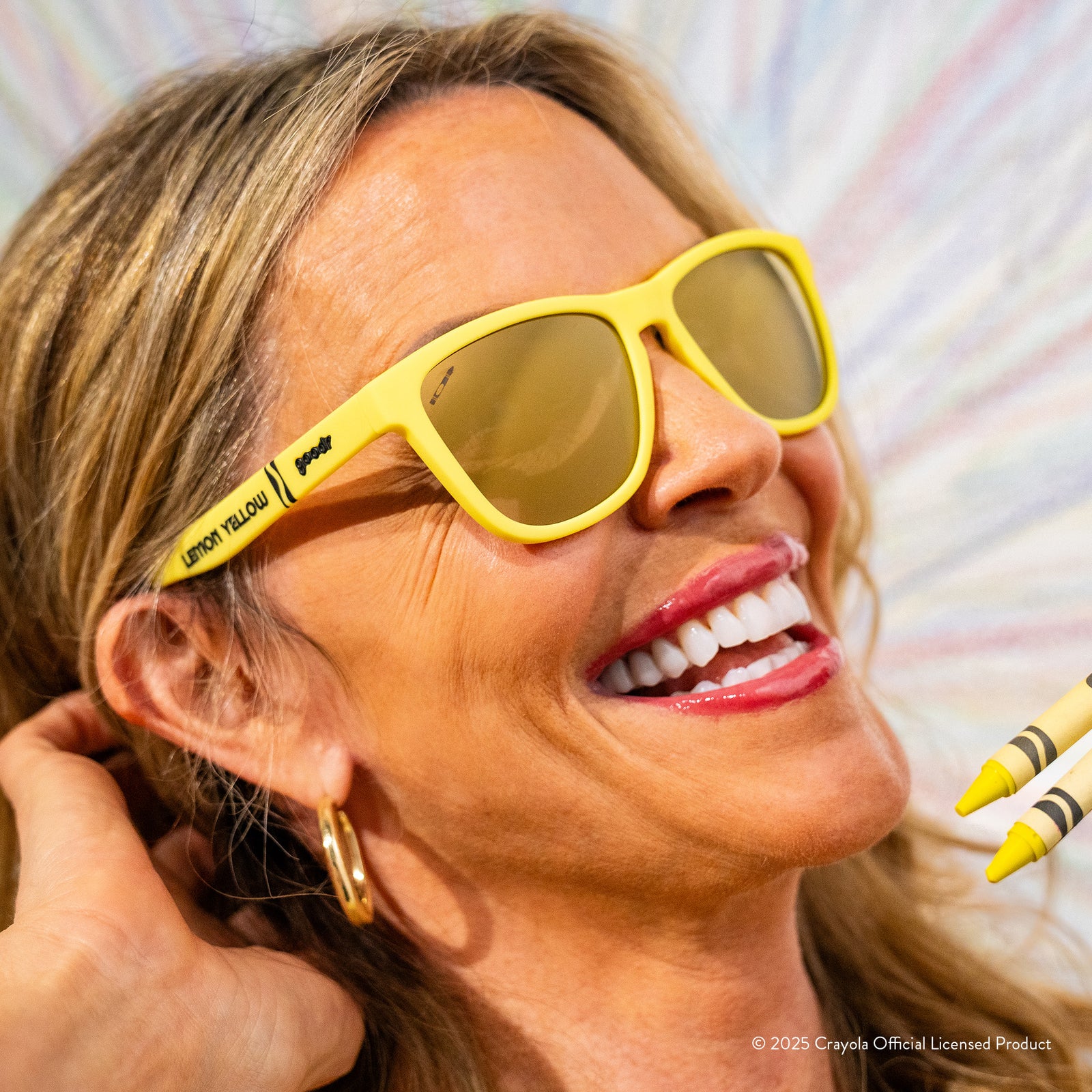 A smiling blond woman wearing Lemon Yellow-colored Crayola sunglasses in front of a wall with drawings