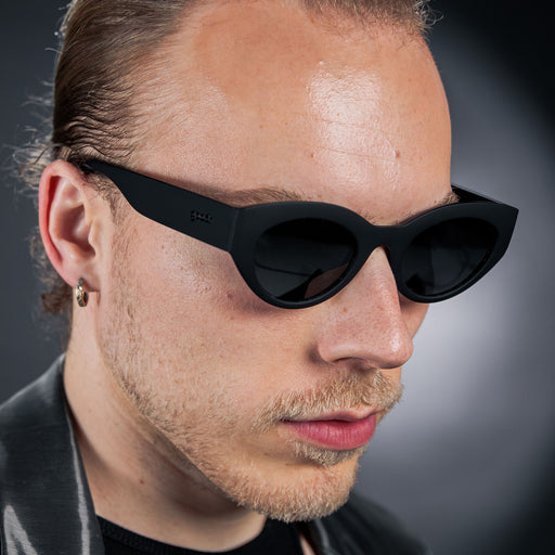 Close-up of a man’s right side profile wearing translucent black, retro-inspired angled cat-eye sunglasses with non-reflective lenses.
