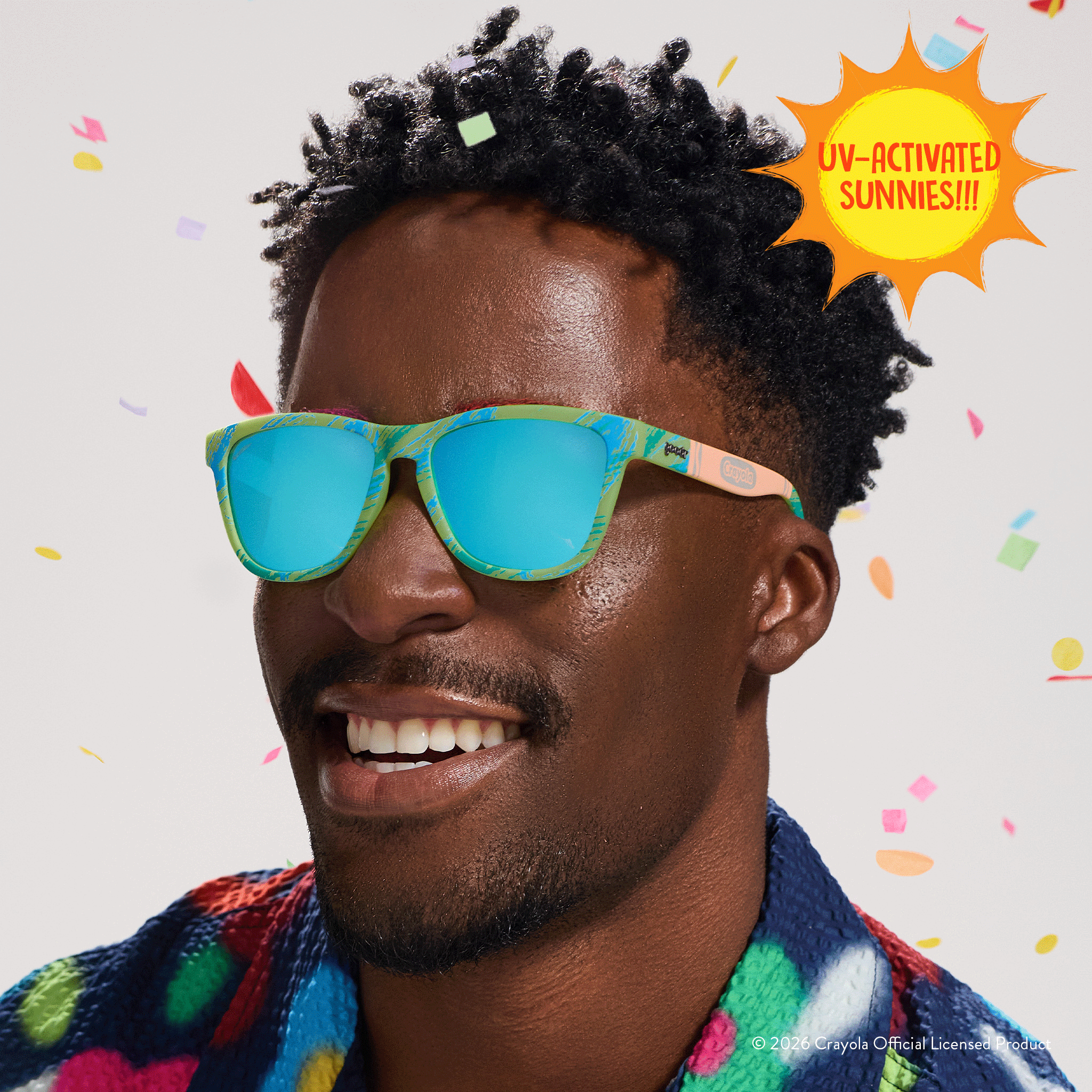 Young man smiles while wearing Crayola goodr UV-activated Confetti Colors sunglasses with light blue reflective lenses.