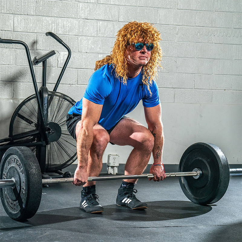 A man in a large curly brown mullet wig and black sunglasses with blue reflective lenses lifts a heavy dumbbell in a gym.