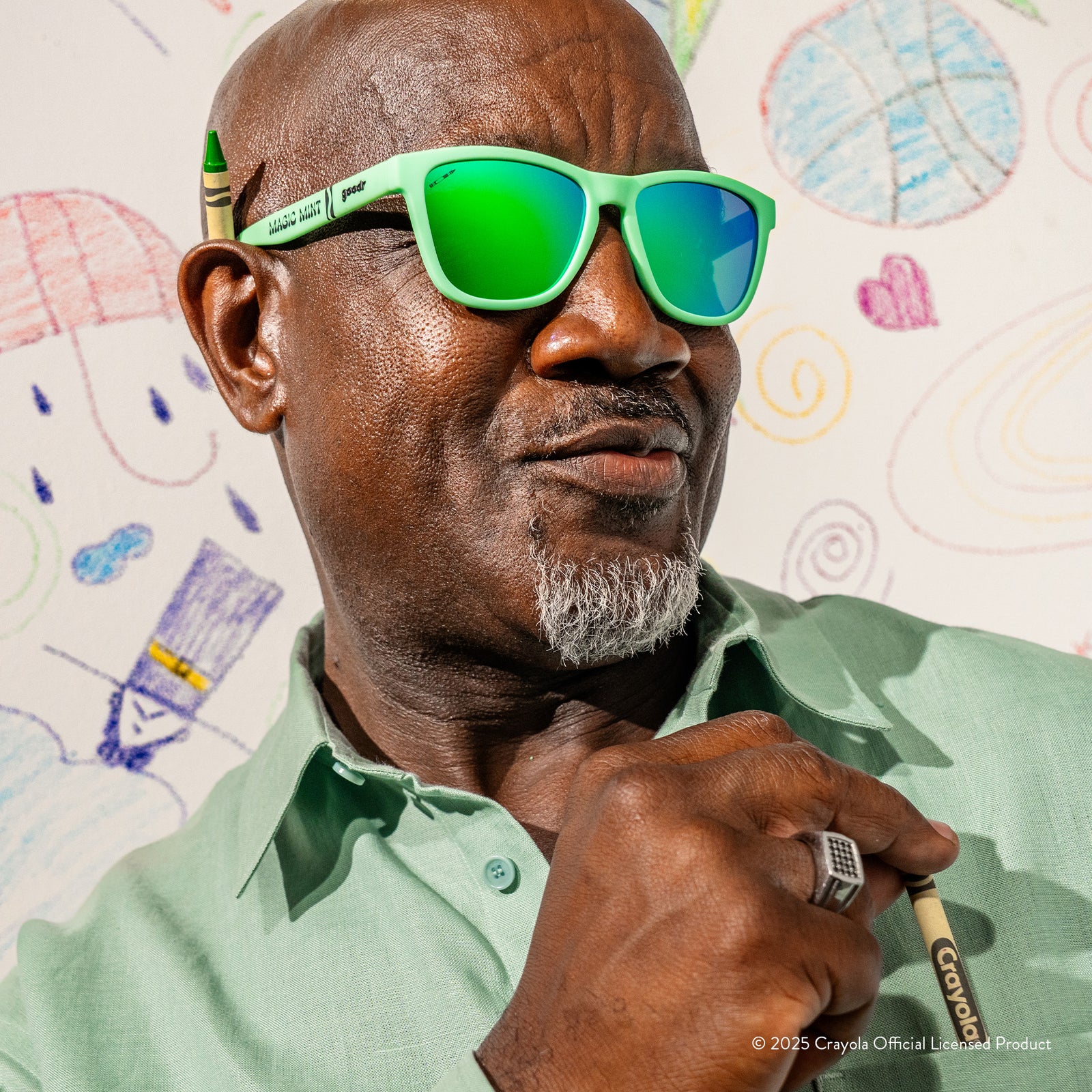 A smirking older man wearing Crayola Magic Mint sunglasses with a crayon under his ear in front of a wall with drawings