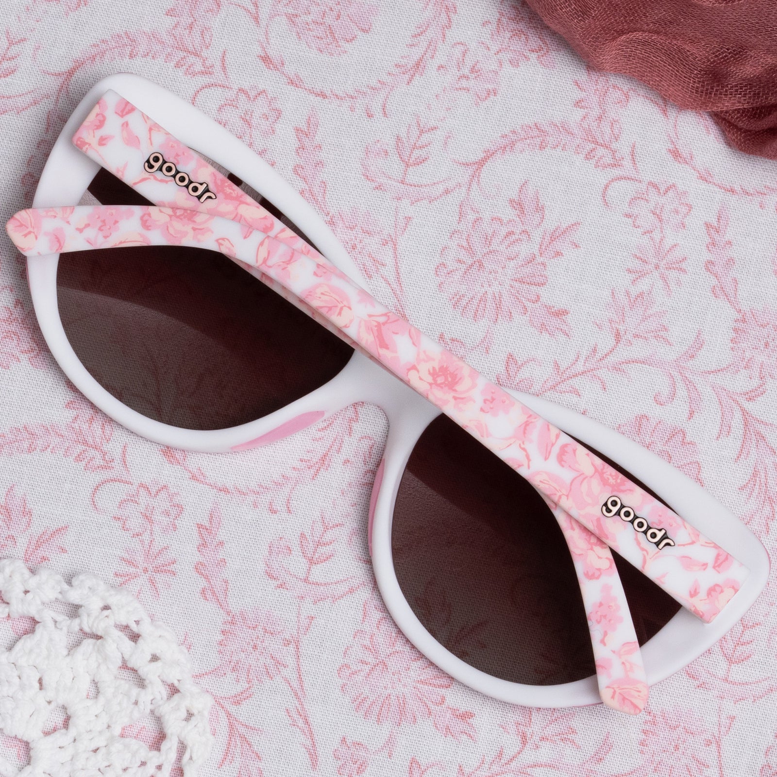 Back view of folded pink and white sunglasses featuring a floral toile pattern and rose gradient lenses on top of a pink floral tablecloth.