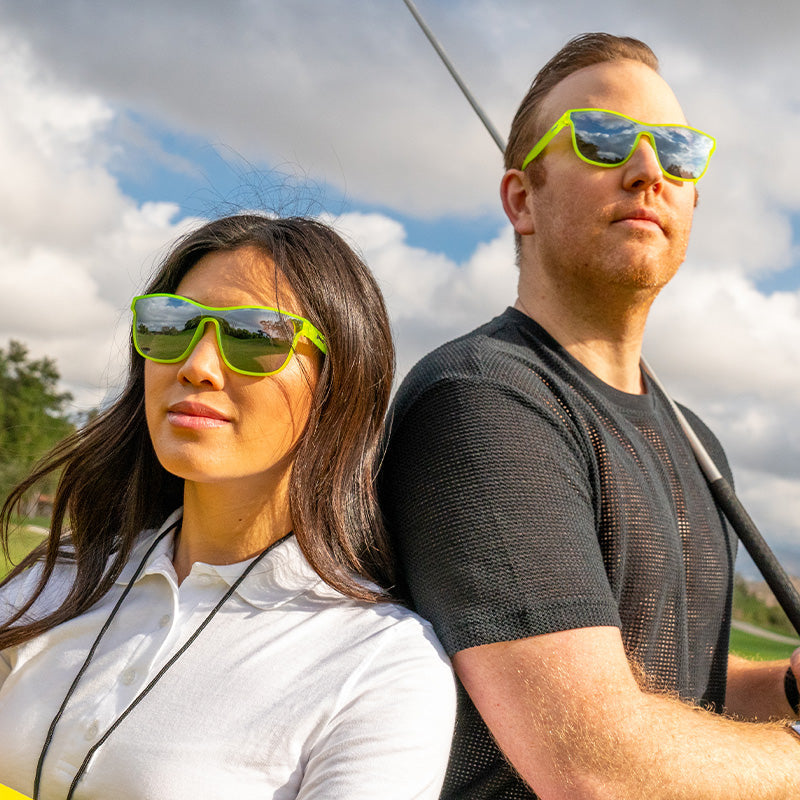 A man and women wearing futuristic square neon green sunglasses with a single mirrored chrome lens staring off into the clouds.