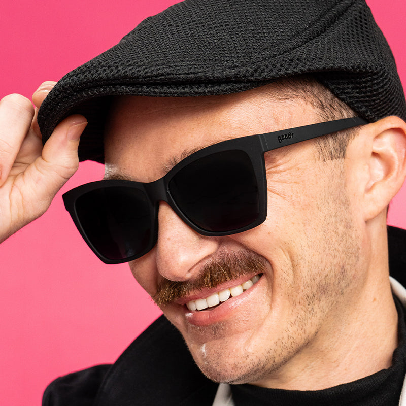 A man in a stylish black outfit with black angled cat-eye sunglasses with non-reflective black gradient lenses smiles.