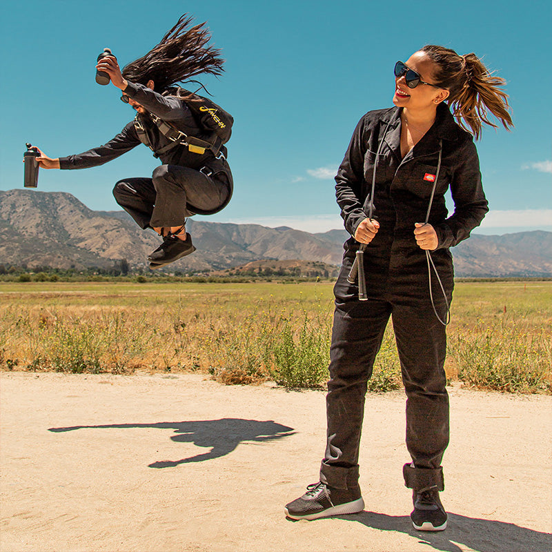 A parachuter double fisting protein shakes lands in a field near a black jumpsuited woman, both wear black aviator sunglases.
