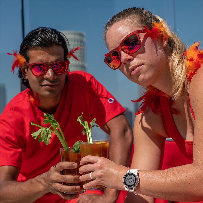A man and woman with orange and red feathers and red sunglasses with red reflective lenses hold bloody marys on a rooftop.