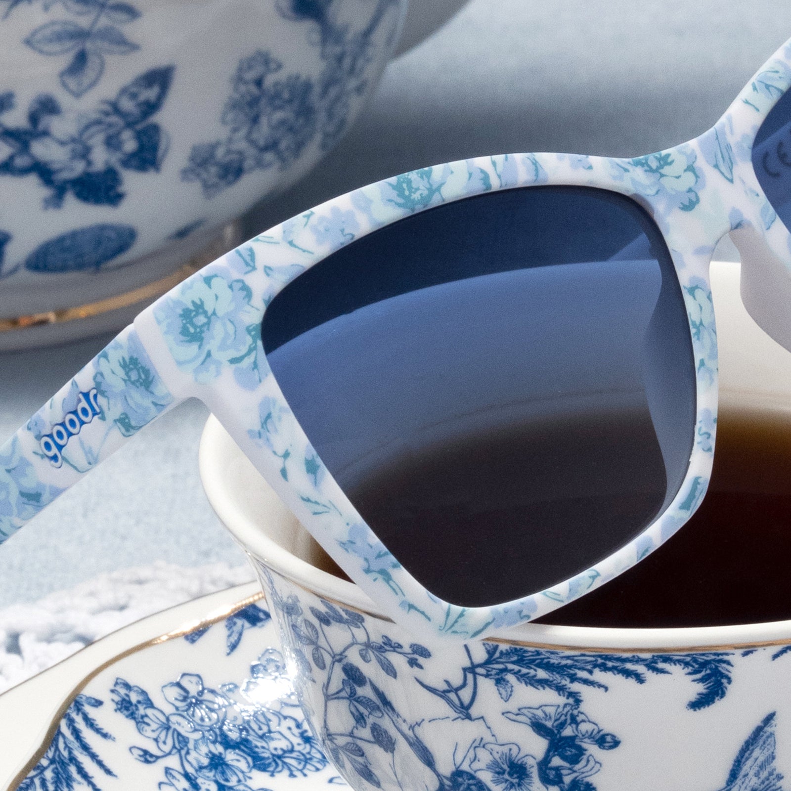 Close-up of blue and white angular cat-eye sunglasses featuring blue gradient lenses on top of a tea-filled teacup.