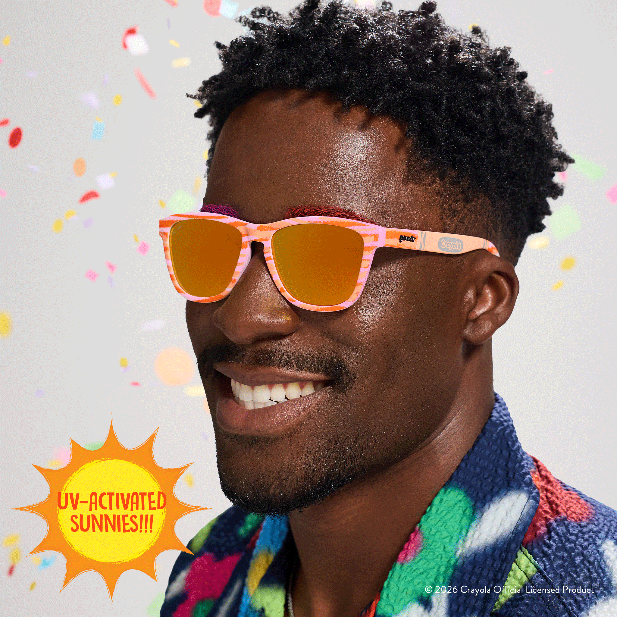 Young man smiles, wearing Crayola goodr UV-activated sunglasses with bone-to-magenta frames and amber reflective lenses.