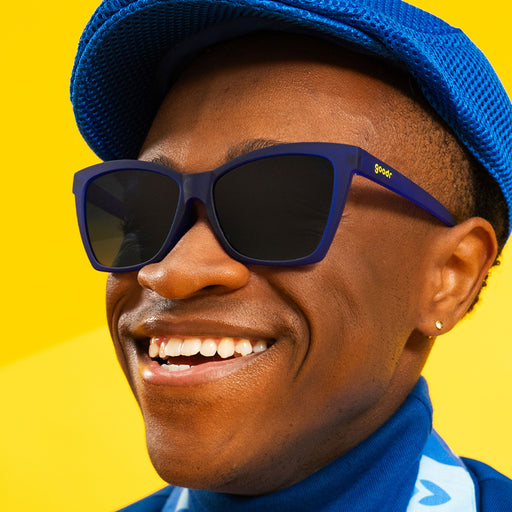 A man in a stylish blue outfit with dark blue angled cat-eye sunglasses with dark blue gradient lenses smiles.