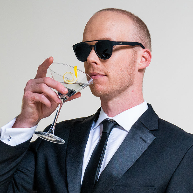 A man wearing a tuxedo and round black sunglasses with a nose bridge & round black lenses sips a martini with a lemon twist.