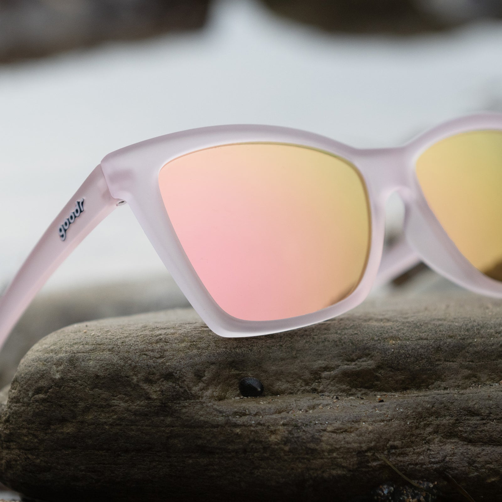Close-up of translucent pink angular cat-eye Pop Gs with light pink reflective lenses propped on a gray seaside rock.