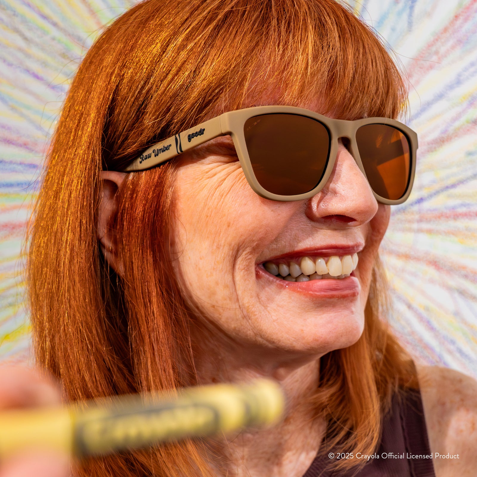 A smiling redheaded woman in a fedora wearing Raw Umber-colored Crayola sunglasses in front of a wall with drawings