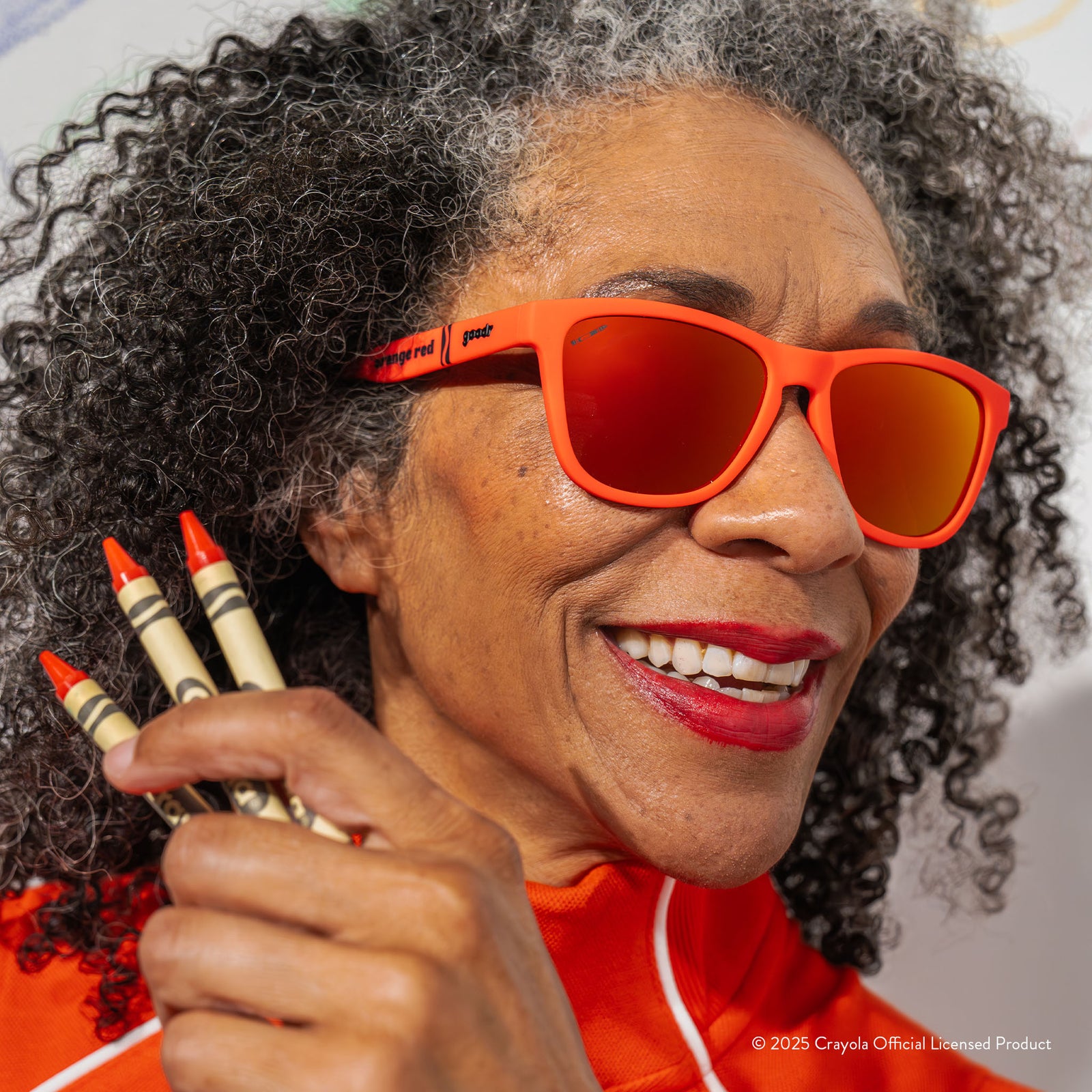 An smiling older woman wears Orange Red Crayola sunglasses and holds up three Orange Red crayons