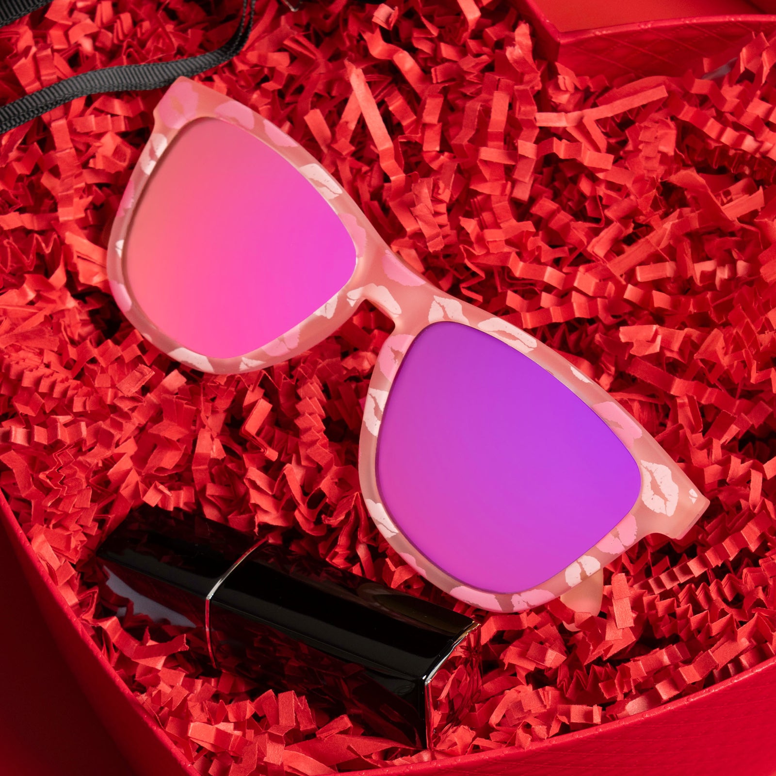 A pair of pale pink sunnies with rose reflective lenses and flirty all-over-kiss print sits in a gift box filled with red crinkle-cut shredded paper.