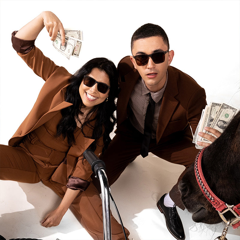 A man and woman in brown tortoiseshell sunglasses with brown non-reflective lenses and brown suits flash fistfuls of cash.