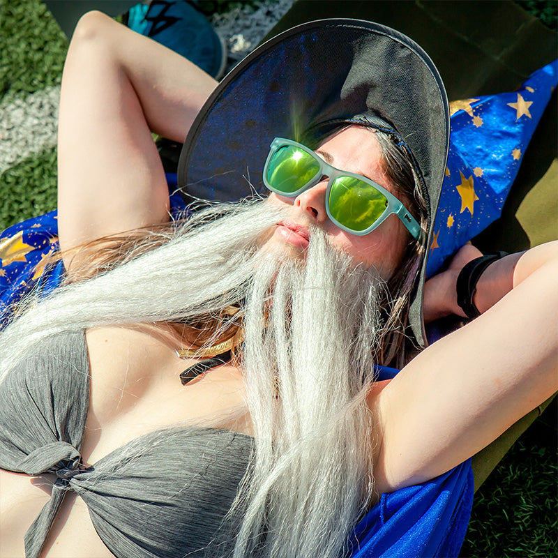 A woman in a bikini with a wizard hat & fake beard sunbathes on a track field wearing light blue sunglasses with gold lenses.
