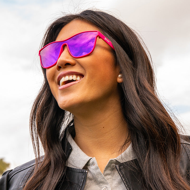 A woman wearing futuristic hot pink sunglasses with a flat purple lens smiles into the distance.